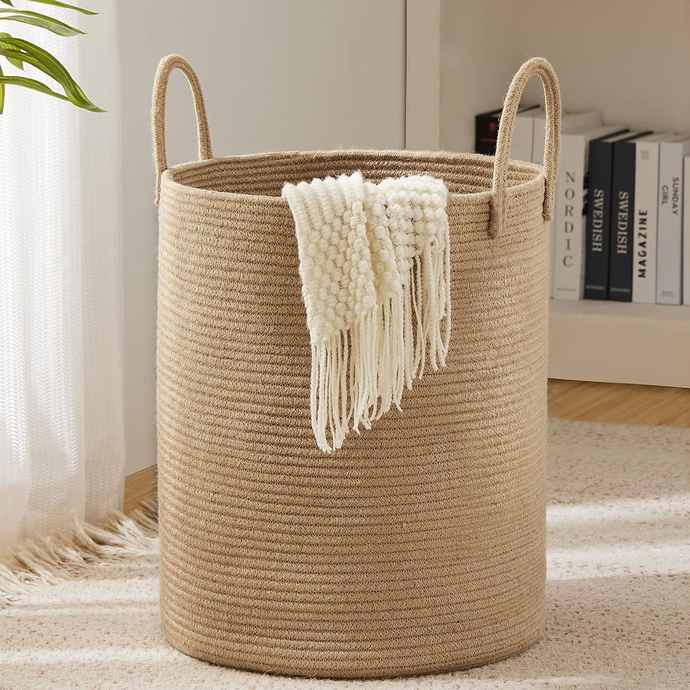 Beige woven basket with handles holds a white textured blanket. It's in a cozy room with books and a plant. Soft, neutral tones.