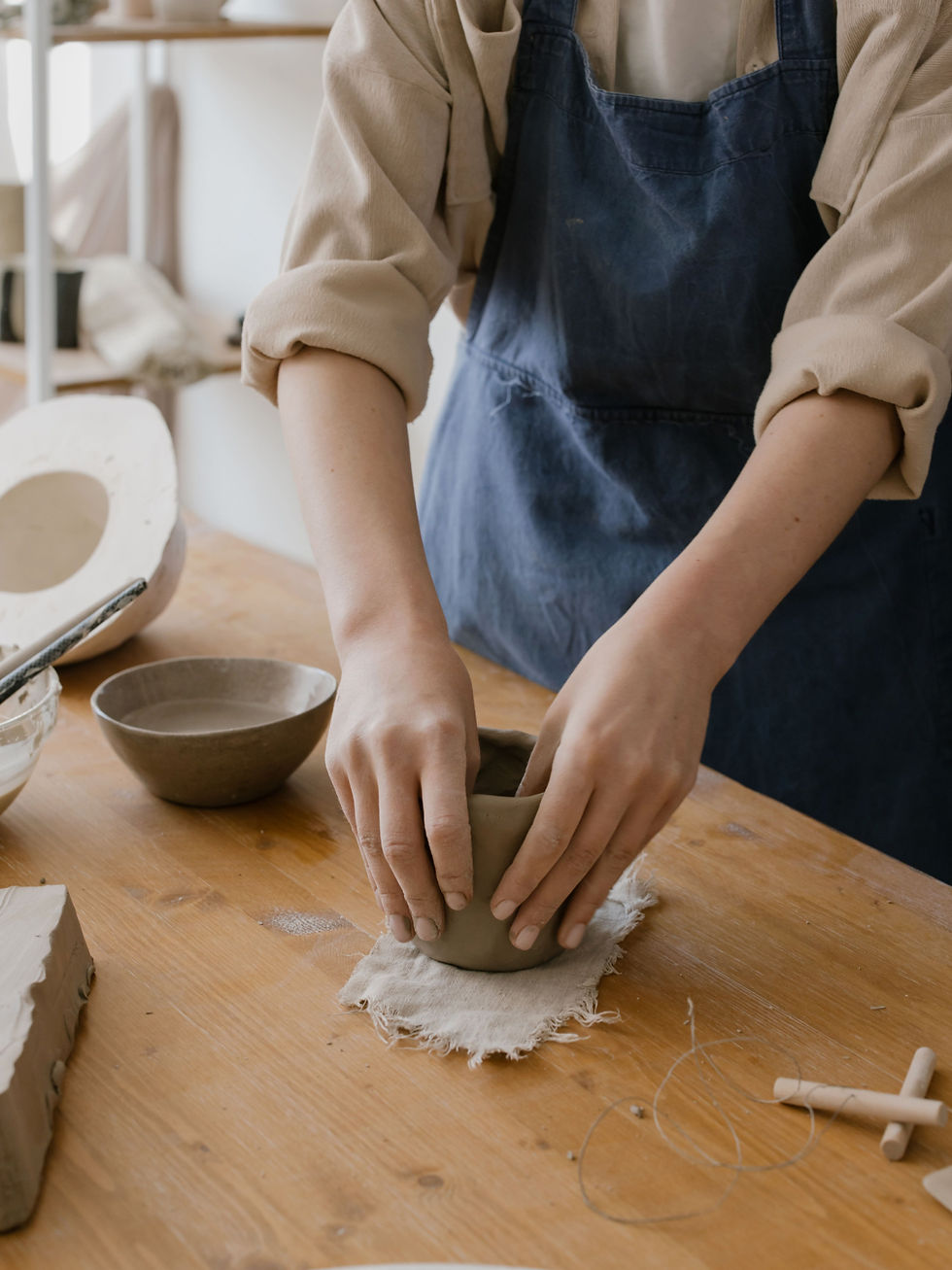 Person in a blue apron shapes a clay bowl on a wooden table in a pottery studio, with tools and ceramics nearby.