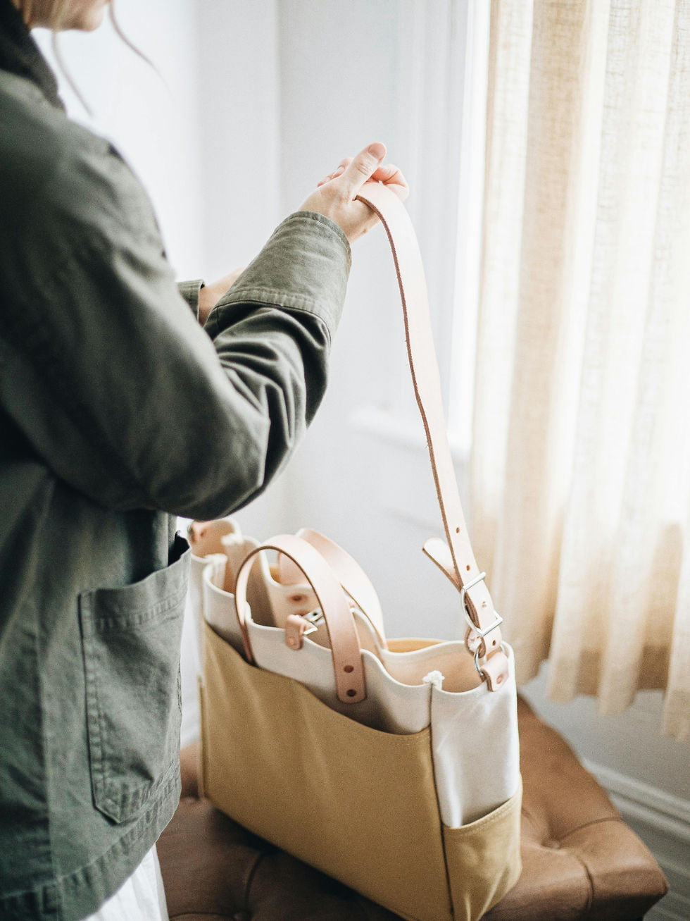 Person in green jacket handling a beige and cream tote bag with leather straps. Soft light filters through a curtain in a cozy room.