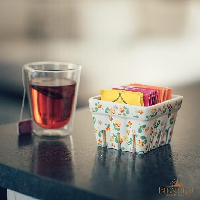 Glass of red tea on a countertop, next to a floral-patterned holder filled with colorful tea packets. Cozy and inviting atmosphere.