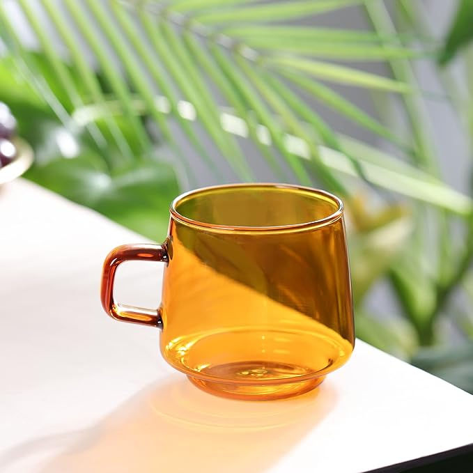 Amber glass mug on a white table with green plant leaves in the blurred background, creating a fresh, minimalist setting.