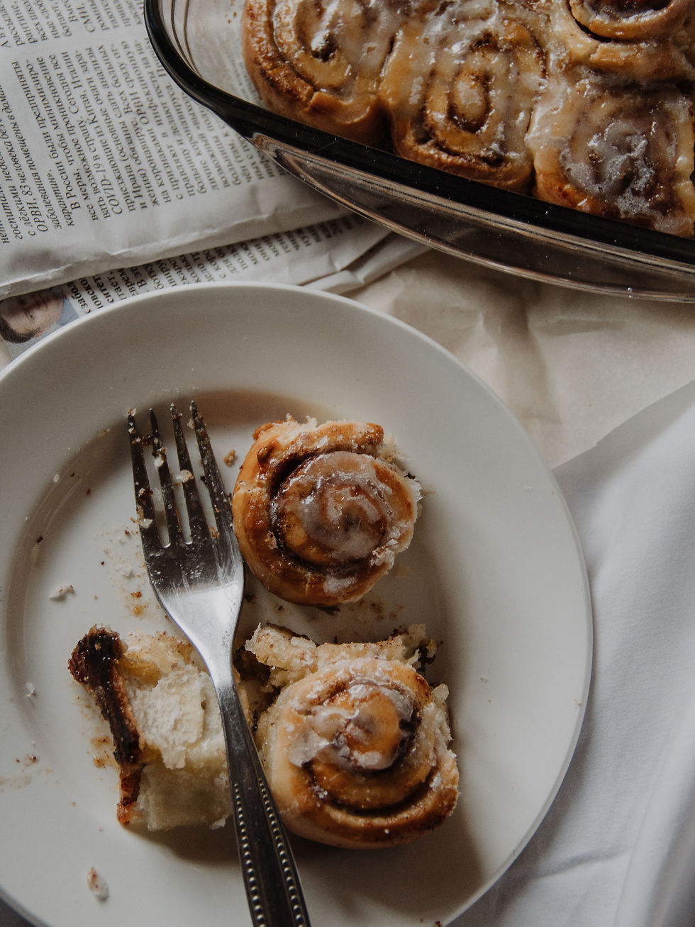 Partially eaten glazed cinnamon rolls on a white plate with a fork, beside a baking dish and newspaper on a beige tablecloth