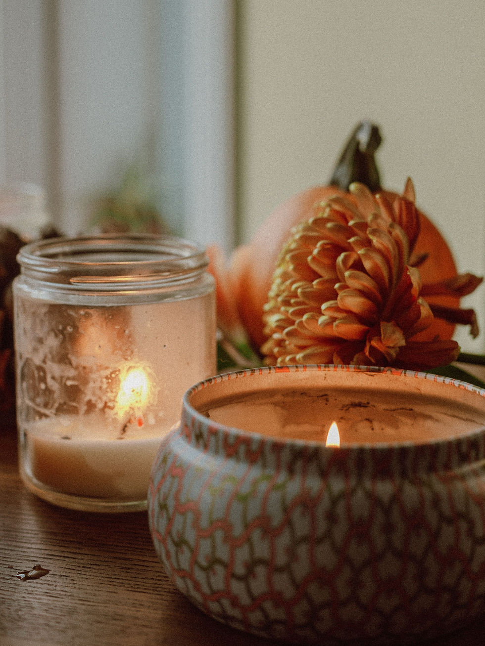 Lit candles in a glass jar and patterned bowl beside an orange flower, creating a cozy autumn mood on a wooden table.