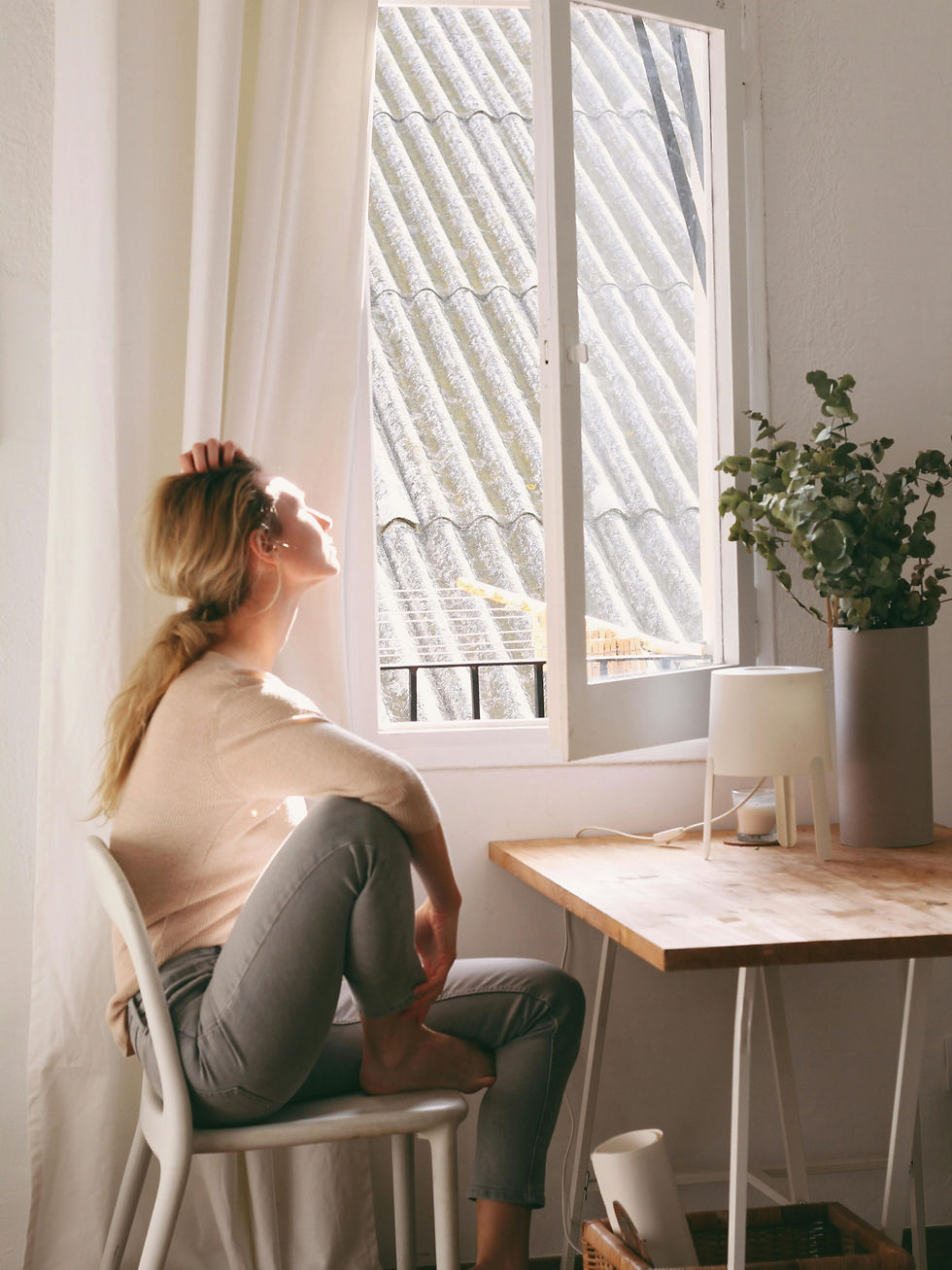 Woman sitting on chair, gazing outside through a window. Sunlight illuminates her face. Indoor plants and lamp on wooden desk nearby. Calm mood.