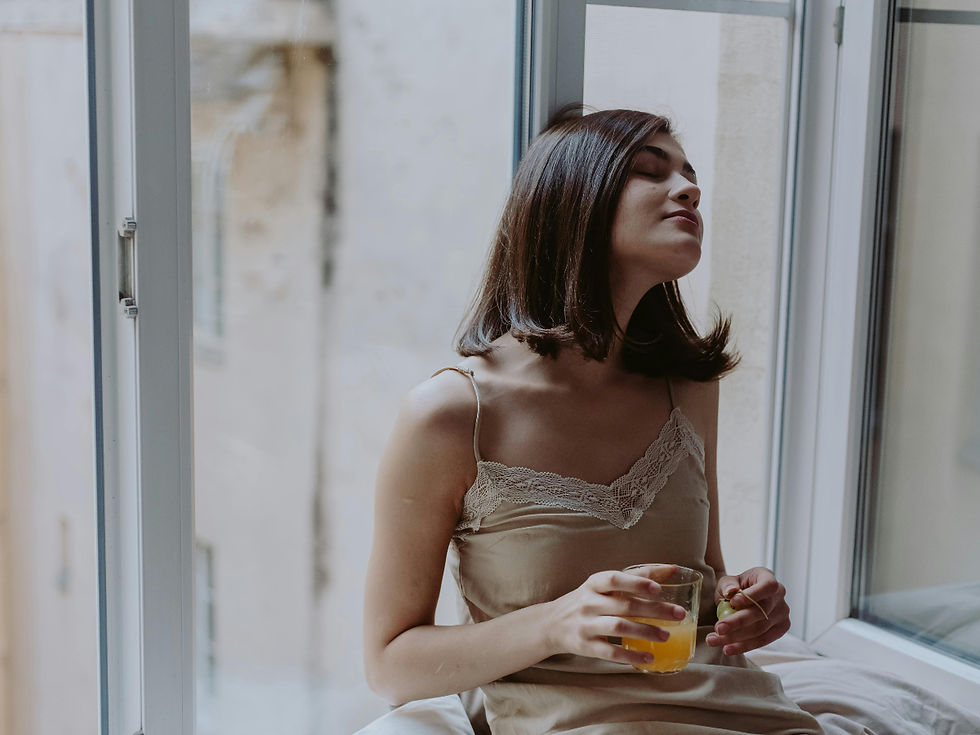 Woman in beige dress relaxes by an open window, eyes closed, holding a glass of orange juice, exuding a peaceful, serene mood.