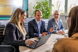 Four colleagues sit around a conference table while one person holds a large caliper during a technical discussion, with a presentation screen in the background reading “streamlining and simplifying supply chain.”