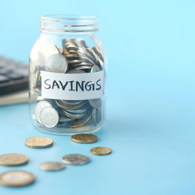 Glass jar labeled “Savings” filled with coins on a blue background, with scattered coins and a calculator in the background, representing how to build an emergency fund and personal financial planning.