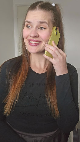 Hymyilevä nainen, jolla on pitkät, ruskeat ja latvoista vaaleammat hiukset. Hän pitää keltaista älypuhelinta korvallaan puhuessaan ja katsoo iloisena sivulle. Hänellä on nenäkoru ja musta pitkähihainen paita, jossa on kohokuvioitu teksti. Taustalla näkyy vaalea ovi ja seinä. Mirva Rontti Creates.