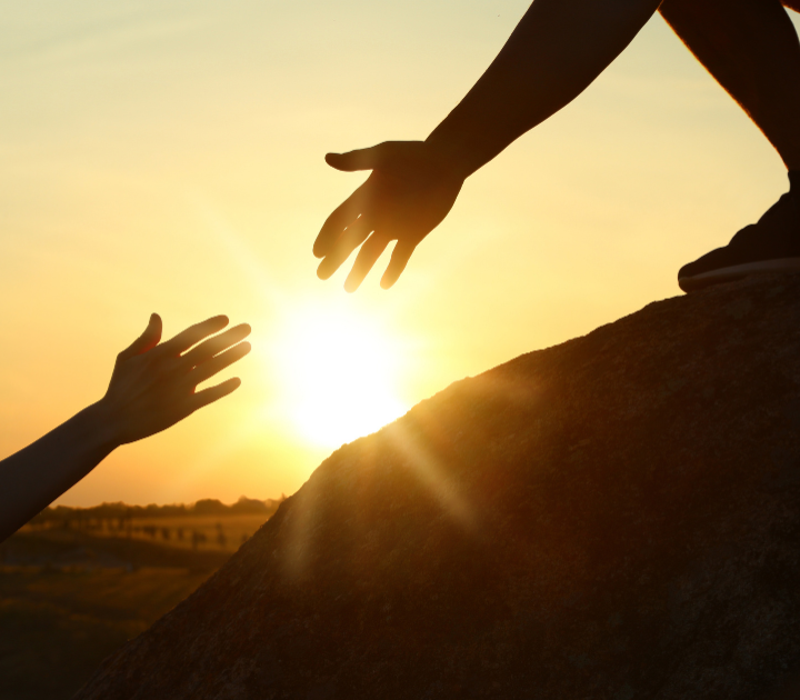 A picture of someone reaching out to help another person as the sun sets in the background.