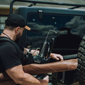Man adjusting equipment attached to a black Jeep's rear, with spare tire. Mbogo Media.
