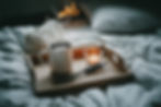 A warm ritual scene with a mug of herbal tea on a wooden tray beside a lit candle, set on a soft bed — creating a cozy, grounding atmosphere for a morning or evening wellness ritual.