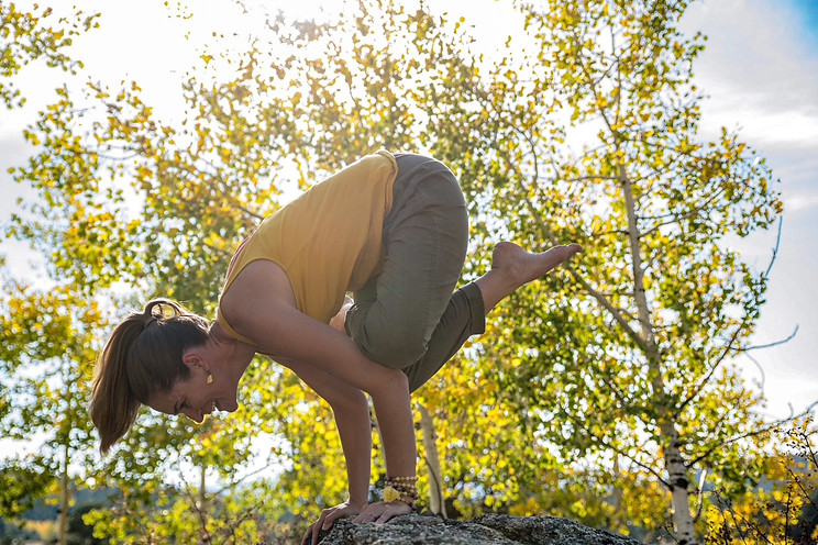 Mindy Arbuckle master yoga teacher in crow pose on top of a rock with glowing trees in the background