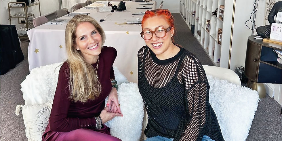 Minqwa and Natasha smiling while seated indoors before The Higher Connection workshop. Behind them is a long table with white chairs, gold star decorations, and shelves, creating a warm and inviting atmosphere