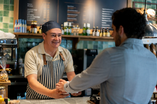 Smiling barista in striped apron shakes hands with a customer at a cafe counter, with menu boards and bottles behind.