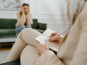Therapist in beige suit takes notes in a notebook while a blurred client sits on a green sofa in a calm counseling room.