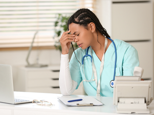 Enfermera en uniforme azul, sentada en un escritorio blanco, con gesto de cansancio. Hay una laptop, papeles y un fonendoscopio.