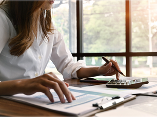 Woman in a white shirt reviews papers, using a calculator and pen at a desk by a sunny window.