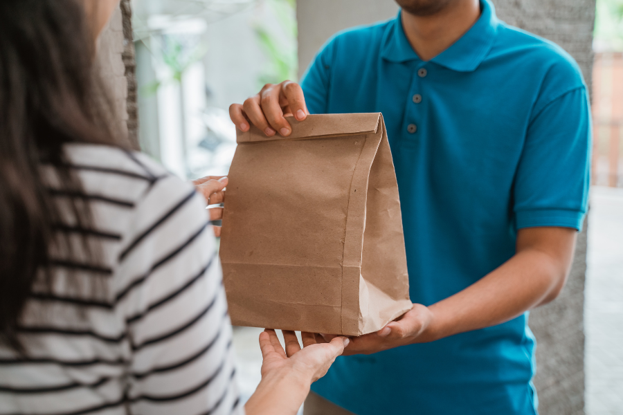 Delivery worker in a blue polo hands a brown paper bag to a woman in a striped shirt at a doorway.