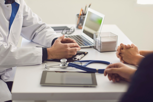 Doctor talks with patient at a desk with a laptop and stethoscope in a bright clinic office.