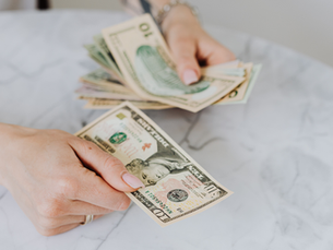 Manos contando billetes de dólar en una mesa de mármol blanco; se ven billetes de 10 y 20 dólares.