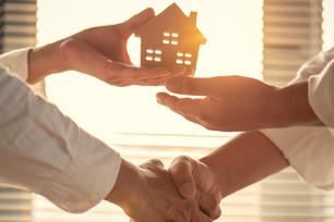 Two people shake hands while holding a small house model in bright sunlight by window blinds, suggesting a real estate deal.