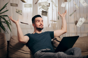 Man on sofa with laptop tosses money into the air, creating a joyful scene. Neutral-toned room with a plant in the background.
