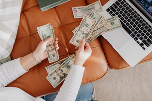Hands counting US dollar bills over a brown couch, next to a laptop. Relaxed setting, partial jeans visible in the background.