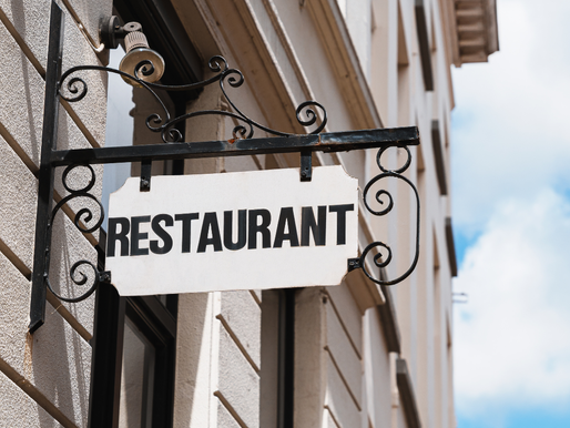 Letrero de "RESTAURANT" en fachada de edificio beige y cielo azul de fondo. Diseño con soporte metálico ornamental.