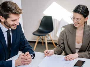 Man in blue suit and woman in glasses review documents at a desk, sharing a smile. Sunlight illuminates the modern office setting.