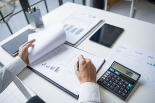 Person reviewing financial charts with a calculator showing 589.7. Papers, tablet, and pen on a white desk in a bright office.