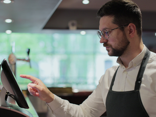 Man in glasses and a black apron taps a touchscreen at a café counter, focused in a softly lit interior.