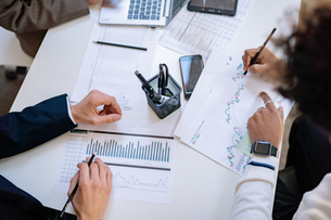 Colleagues review charts and graphs at a white desk, one person marking a growth chart with a pencil beside phones and a laptop.