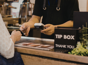 Persona paga con tarjeta a un cajero en cafetería. Caja de propinas negra con "TIP BOX THANK YOU". Ambiente cálido y acogedor.