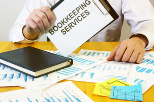 Person in white shirt holds a clipboard labeled BOOKKEEPING SERVICES over financial charts, with a notebook on a desk.
