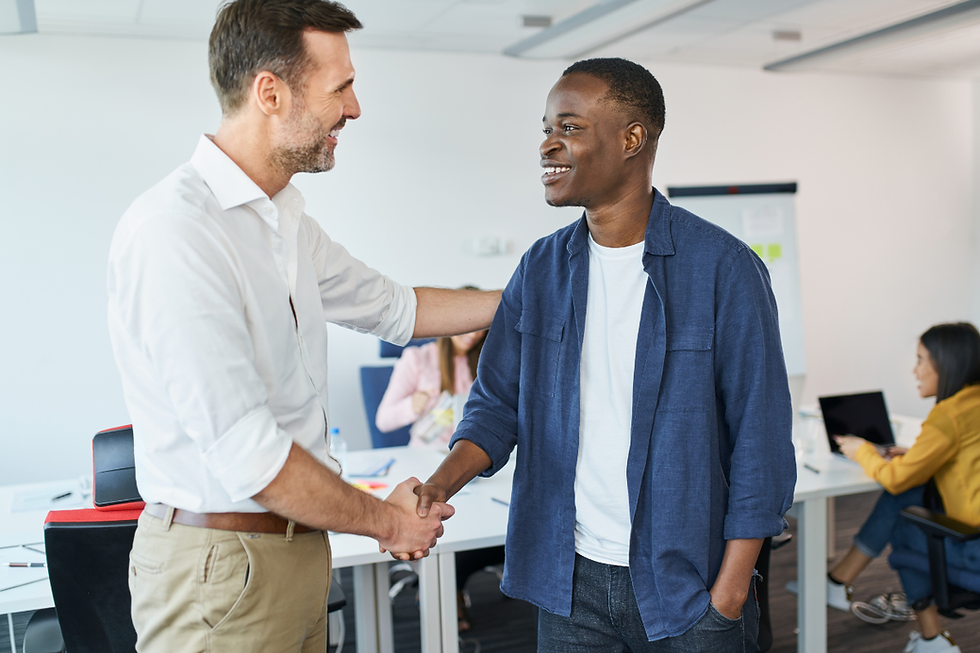 Two men shake hands and smile in a bright office. A woman works at a laptop in the background. Collaborative and friendly mood.