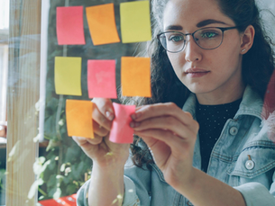 Woman placing pink sticky notes on a glass board indoors. Notes are pink, orange, yellow. She looks focused, wearing glasses and denim jacket.