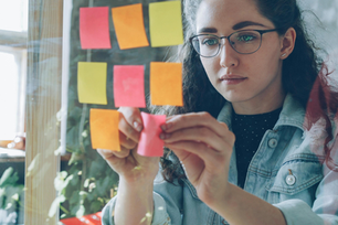 Woman placing pink sticky notes on a glass board indoors. Notes are pink, orange, yellow. She looks focused, wearing glasses and denim jacket.