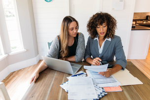 Two women review bills at a wooden table, one using a calculator beside a laptop and paperwork in a bright kitchen.