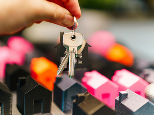 Hand holds house-shaped keychain with keys; blurred background of colorful miniature houses in shades of pink and orange.