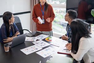 Team meeting in a bright conference room, woman presenting design mockups while colleagues listen around a laptop-covered table.