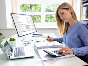 Mujer rubia en oficina luminosa calcula facturas junto a un monitor con Invoice, portátil y calculadora; ambiente concentrado.