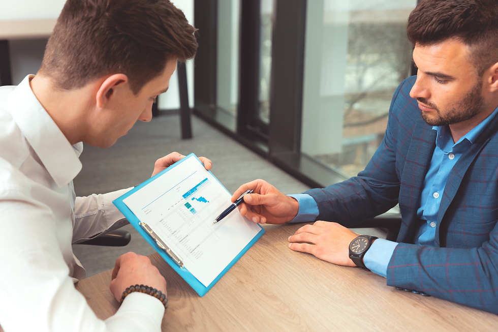Two men in formal attire discuss a clipboard document with charts in an office. One points with a pen. Mood is focused and professional.