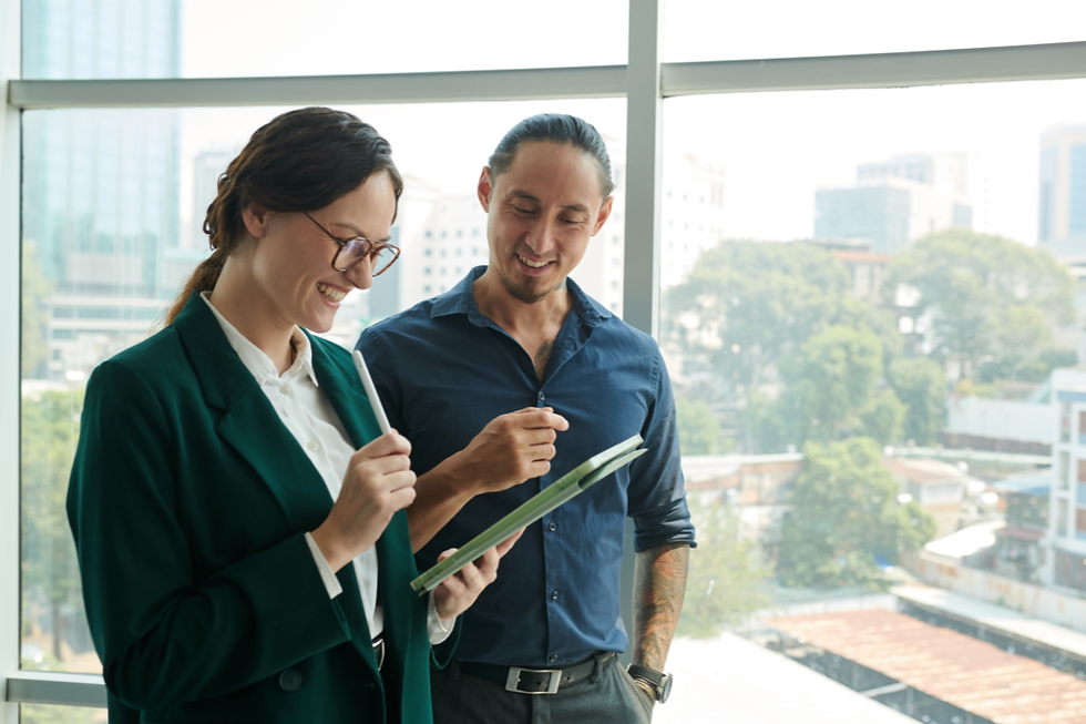 Smiling coworkers in an office by large windows, one holding a tablet and stylus while the other looks on, with city buildings outside.