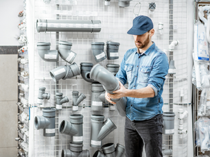 Man in a blue cap inspects a gray plumbing pipe fitting in a hardware store aisle with rows of PVC parts on a white grid wall.