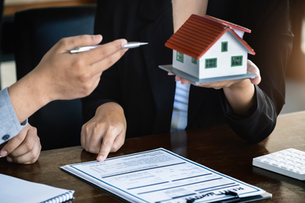 Two people review a real estate contract as one holds a model house and another points with a pen at a form on a desk.