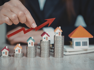 Hand points to red arrow rising over coins with mini houses on top, symbolizing real estate growth. Background features a large house model.