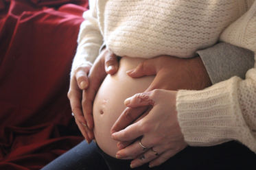 A pregnant woman with a white sweater pulled up over her belly sits and rests her hands on top of another set of hands that are reaching from behind her and placed on her stomach.
