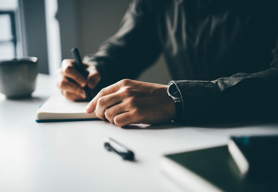 Hands writing in a notebook on a white table with a black pen nearby, a cup in the background, creating a calm, focused mood.