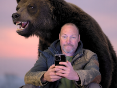 Man taking selfie with a bear in snowy forest, wearing green jacket and backpack. Bear looks alert; forest background is dimly lit.