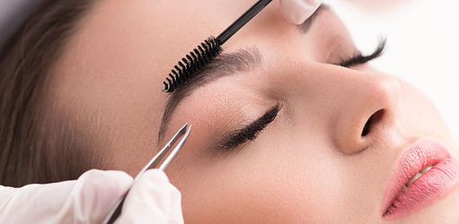 Gros plan sur un visage féminin , yeux fermés, vu de biais, sur lequel on voit un goupillon noir brossant un sourcil d'une main et de l'autre main est tenue une pince à épiler